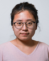 An Asian woman with black hair tied up is wearing black-framed glasses and a pale pink v-neck sweater against a white-grey background.
