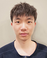 An Asian man in his twenties, with medium length, frizzy black hair. He has a neutral expression and is wearing a navy Henley shirt. Behind him is a white wall.