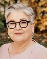 Grey-haired pale-skinned woman smiling, wearing bold dark glasses and silver hoop earrings. Wearing a pink round necked shirt. Background is blurred, but of fall changing leaves.
