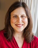 Laurie Proulx has shoulder-length brown hair that is mostly straight. She is a woman with light beige skin and there is a slight scar on her throat where she had a tracheostomy. She is smiling, and the backdrop is her living room, where she sits next to a sunny window.