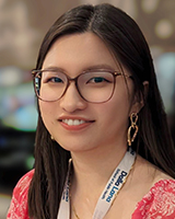 An Asian woman in her mid-twenties, with straight, dark brown hair that falls below the shoulders, and dark brown eyes. She is smiling and wearing rectangular brown glasses with round edges, gold hoop earrings, a thin gold chain necklace, a Dalla Lana School of Public Health lanyard, and a bright pink blouse with white flowers. The background blurred, showing different shades of brown.