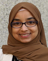 A woman with dark brown eyes is smiling. She is wearing black-framed glasses, a brown headscarf, a black shirt, a white blazer, and two gold necklaces. The white and grey patterned background is blurred.