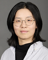 An Asian woman has shoulder-length black hair, thin silver-framed glasses, and a silver nose ring. She wears a black turtleneck and a white shirt with light grey vertical stripes. The background is dark grey.