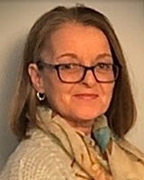A light brown-haired, pale-skinned woman is smiling. She is wearing bold dark glasses and small hoop earrings, along with a white knitted cardigan and a beige scarf featuring light blue, brown, and yellow patterns. The background is white-grey.