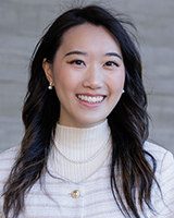 An Asian woman with dark brown eyes and long, wavy black hair with brown highlights is smiling. She is wearing a white sweater along with pearl earrings and a pearl necklace. The background is blurred in beige and light brown.